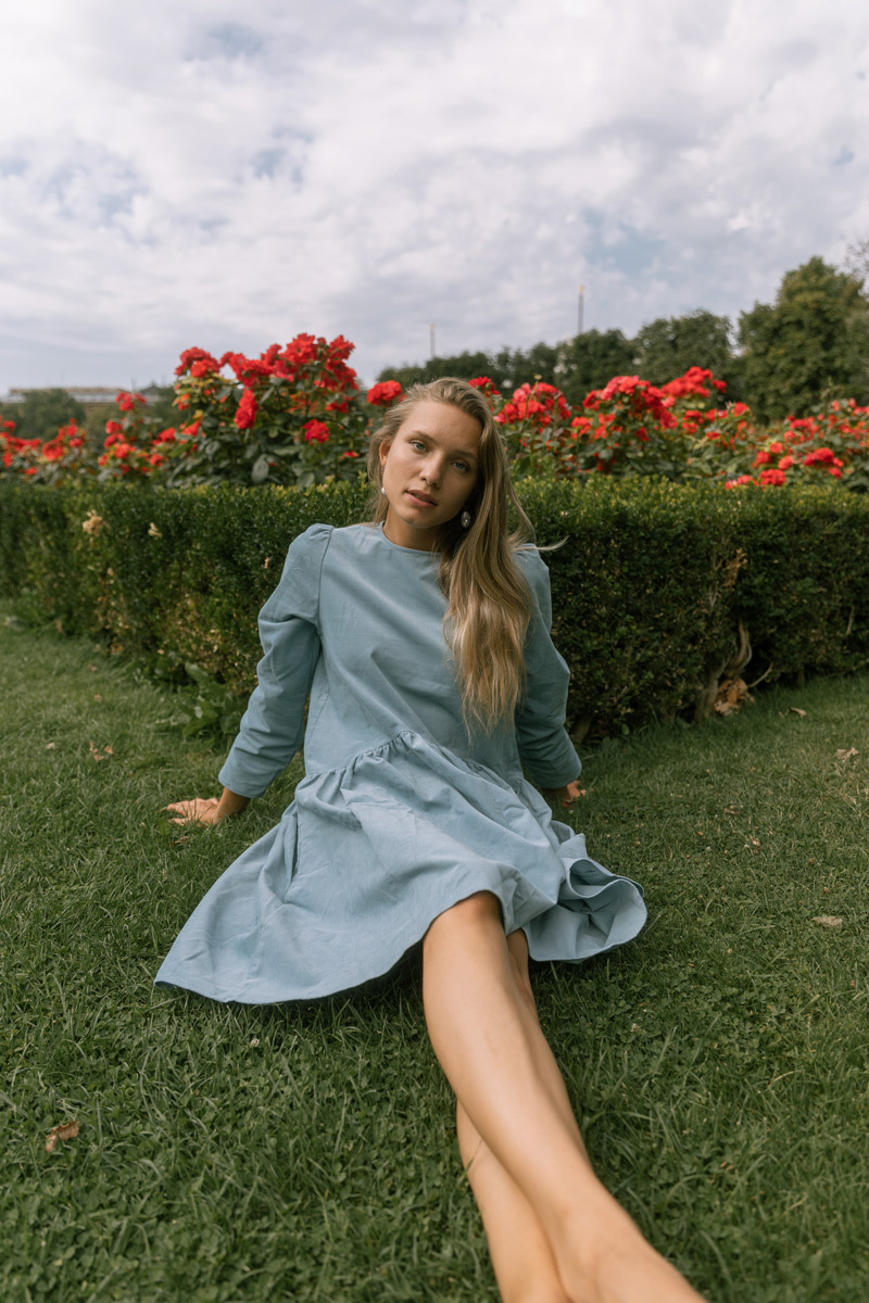 Blonde girl in a blue dress, sitting on the grass. Red roses are behind her