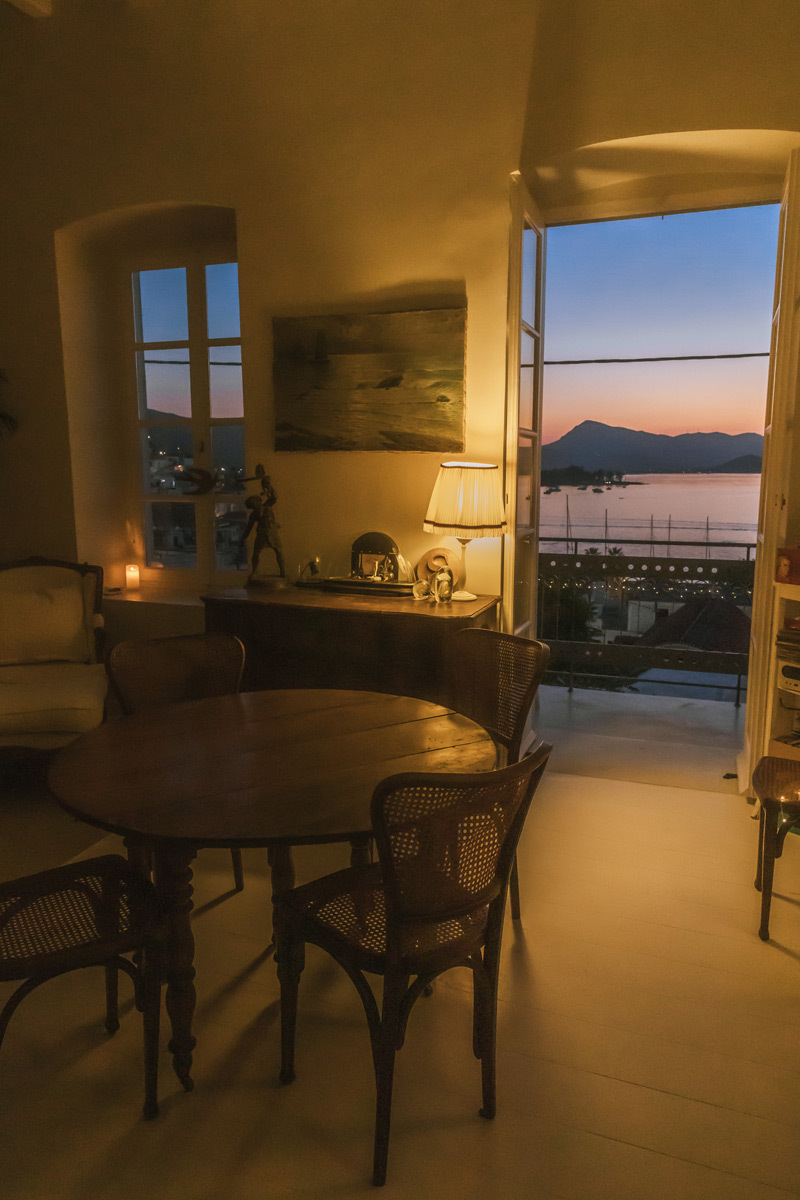 A living room with a big table and chairs. A balkony where you can see the sunset over the sea