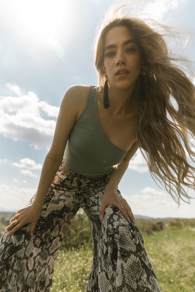 Blonde girl wearing pants and a shirt and leaning towards the camera.