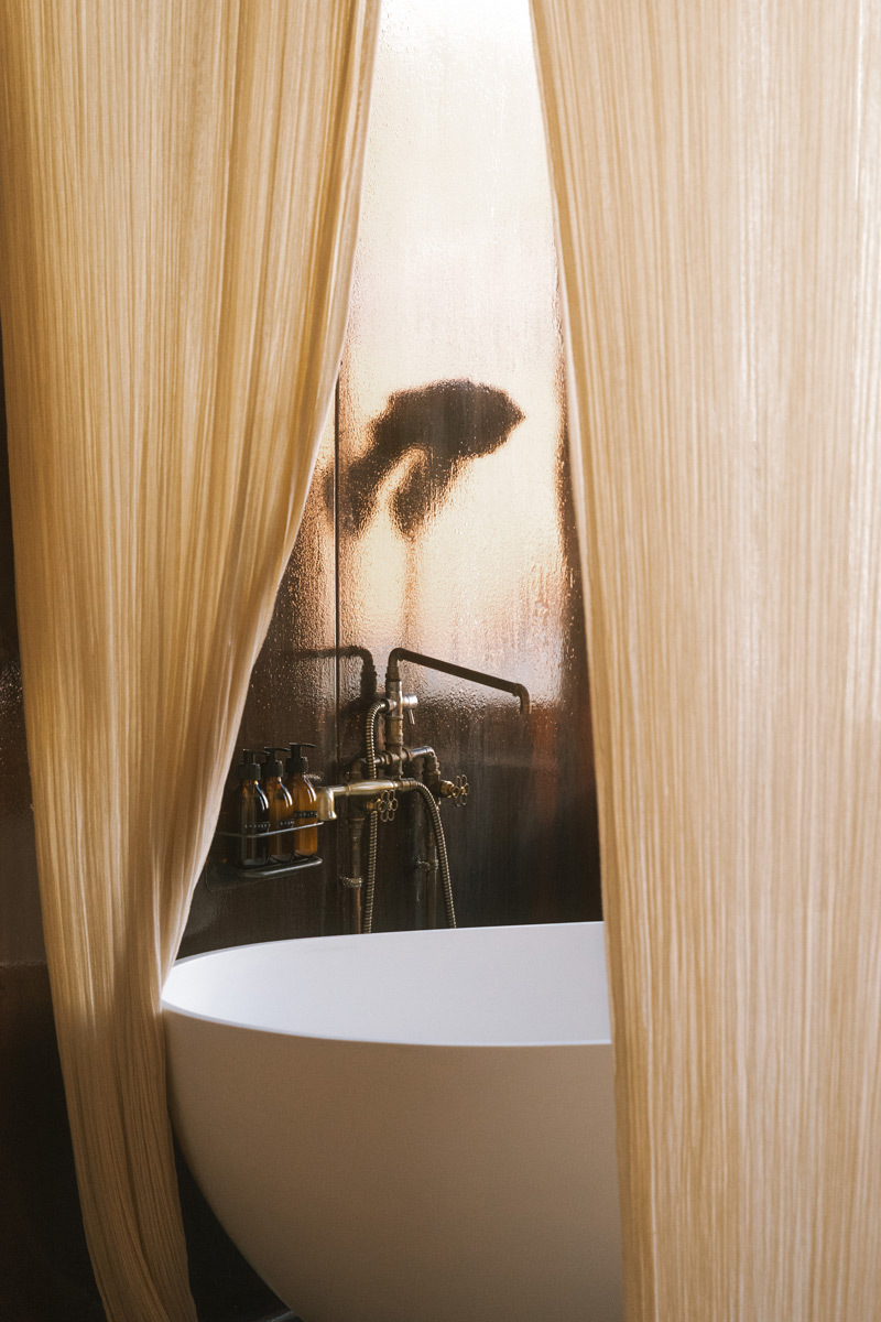 A bathtub that is surrounded by curtains, a reflection of a palm leave on the wall behind of it