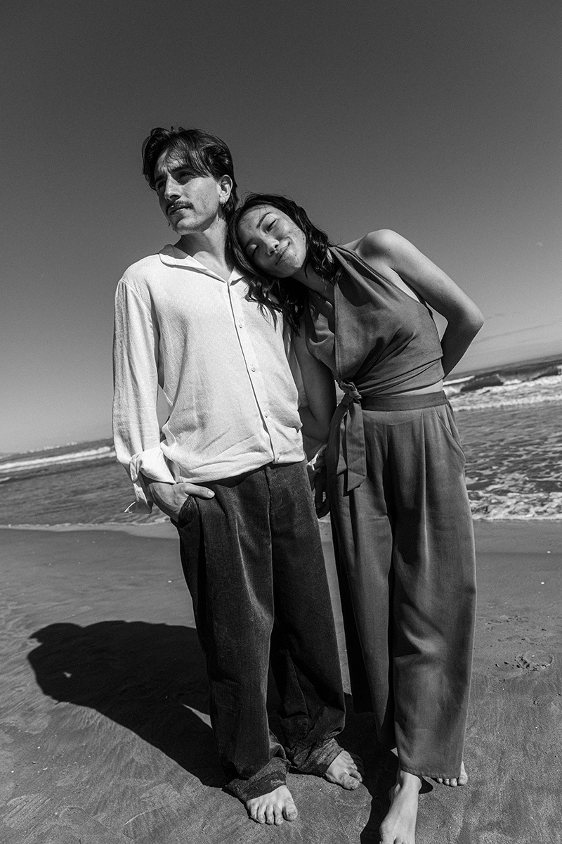 b&w photo of boy & girl standing in front of the ocean, girl leaning head onto boy