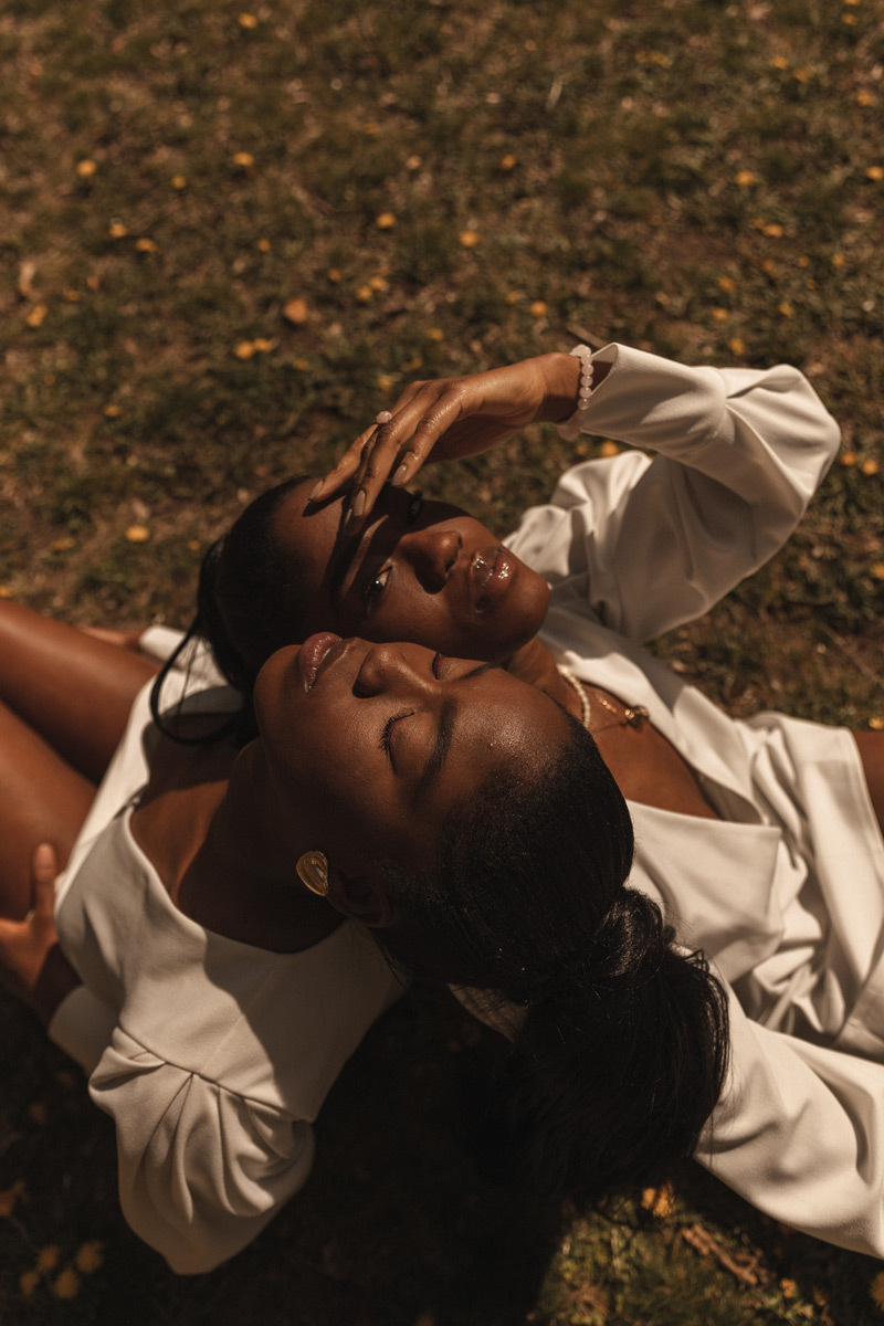 Two twin sisters leaning onto each other while sitting. One has her eyes closed, the other one holds up her hand to have a clear sight despite of direct sun light